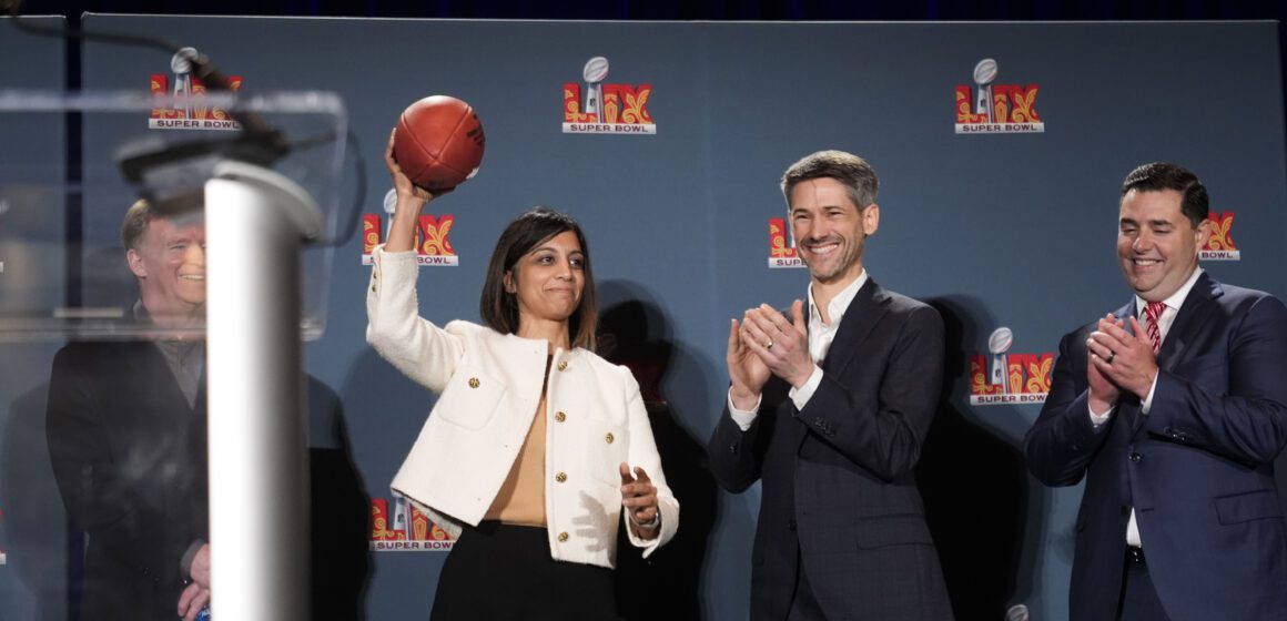 Woman wearing white jacket holding football standing on stage beside applauding men