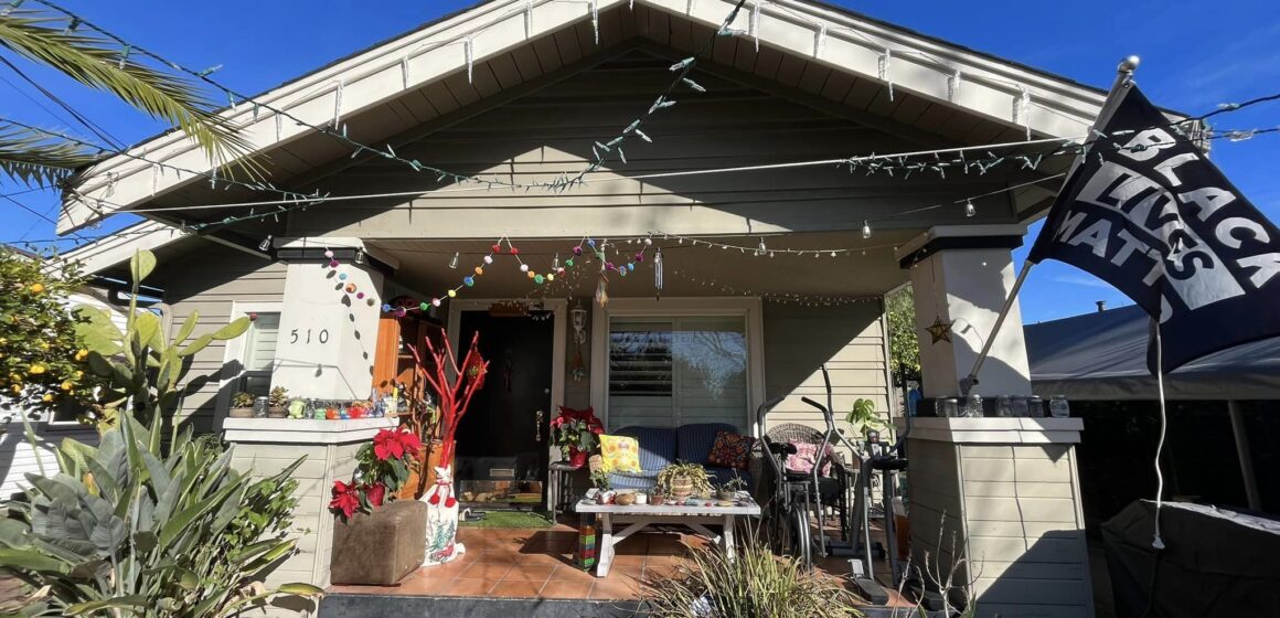 A housefront porch with an exercise bike, dresser filled with shoes, a Black Lives Matter flag and Christmas lights strung up.