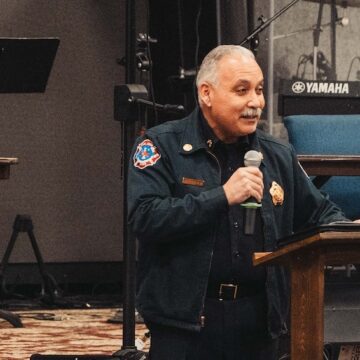 El jefe de bomberos de San José habla ante una multitud en el interior sosteniendo un micrófono.
