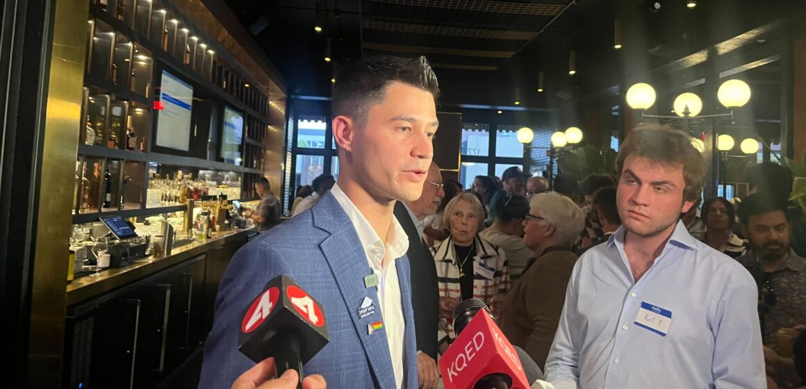 A man in a suit speaking into microphones inside a bar