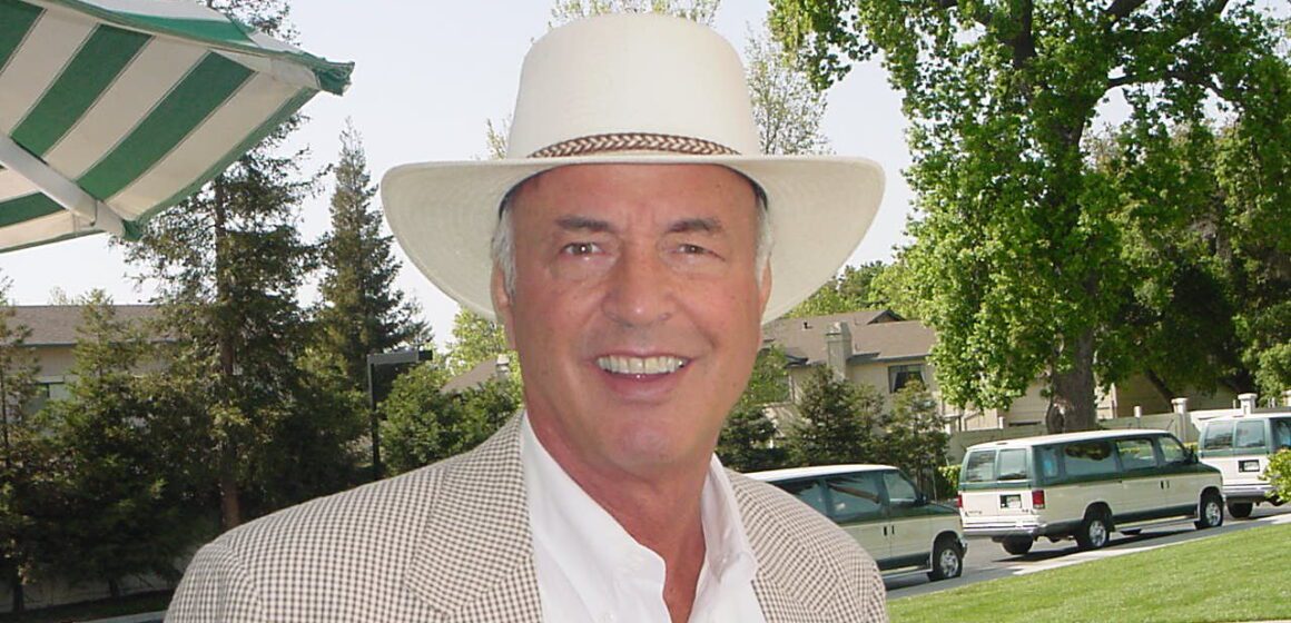 Older light skinned man with white hair smiles wearing a white wide brimmed hat, white button down and checkered blazer