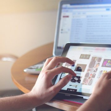 Manos controlando una tableta táctil sobre una mesa, con una computadora portátil en el fondo