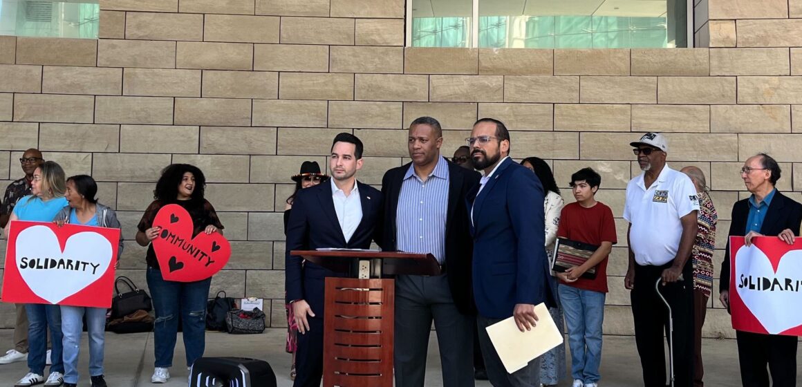 Three men standing at a podium, with a line of people standing behind them