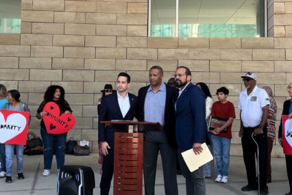 Three men standing at a podium, with a line of people standing behind them