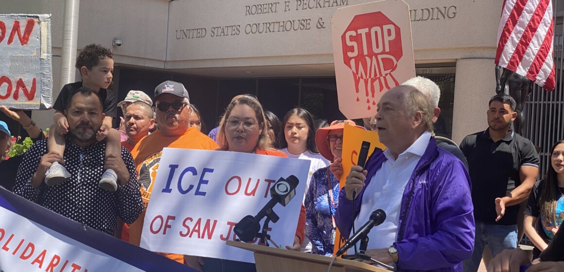 A man speaks into a microphone at a podium with people behind him holding protest signs