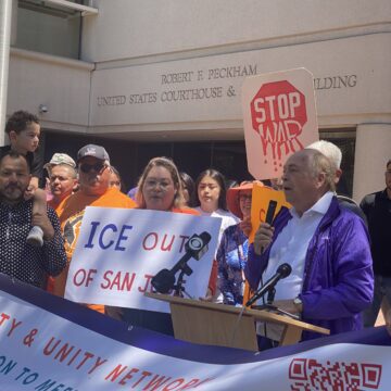 A man speaks into a microphone at a podium with people behind him holding protest signs