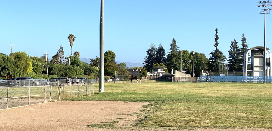 Mostly empty baseball field and lawn with some people standing on side