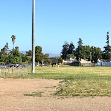 Mostly empty baseball field and lawn with some people standing on side