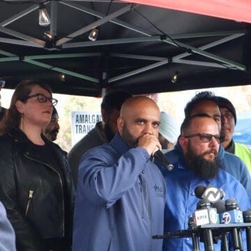 Un hombre calvo de piel oscura que viste un cortavientos azul habla a través de varios micrófonos debajo de una tienda de campaña rodeada de gente.
