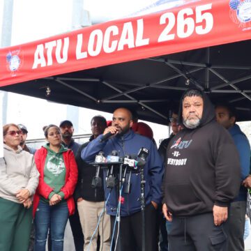 Un hombre calvo de piel oscura, con una cazadora azul, habla a través de varios micrófonos bajo una carpa roja con la inscripción "ATU Local 265", con docenas de personas debajo y carteles pegados a los postes que dicen "Sindicato de Transporte Amalgamado Local 265 en huelga contra VTA".