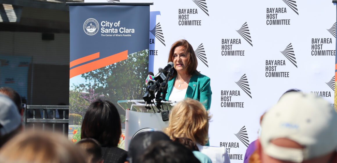 Fair skinned woman with long brunette hair wearing a teal blazer and white shirt speaks at a podium in front of a seated audience, with Bay Area Host Committee and City of Santa Clara banners behind her