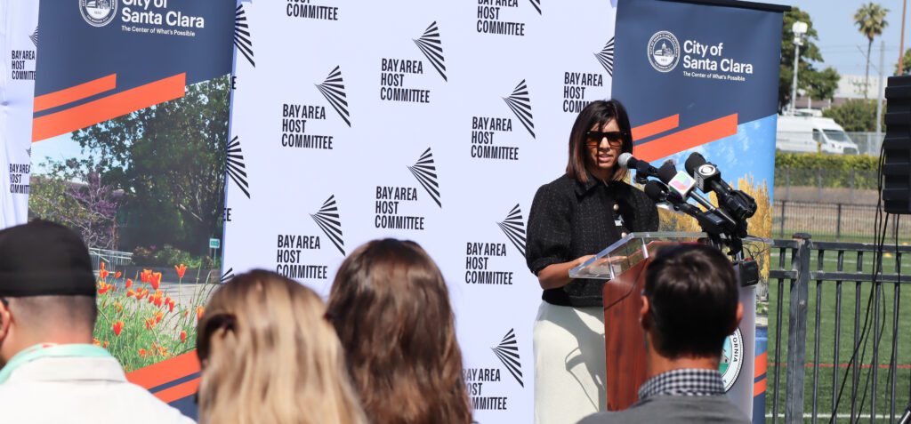 Darker skinned woman with bob wearing black puff sleeved jacket speaks at a podium in front of a crowd, with Bay Area Host Committee and City of Santa Clara banners behind her