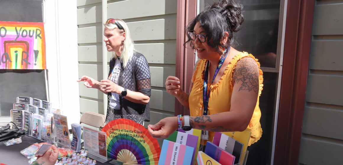 Two women standing behind table at an outdoor venue with open rainbow fan, white and rainbow rubber ducks, multiple resource pamphlets and signs in multiple pride flags