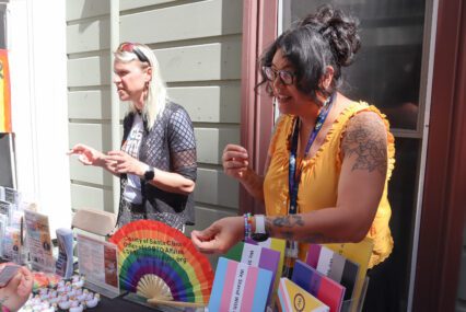 Two women standing behind table at an outdoor venue with open rainbow fan, white and rainbow rubber ducks, multiple resource pamphlets and signs in multiple pride flags
