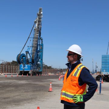 Man in orange high vis vest and a white hardhat watches large construction equipment