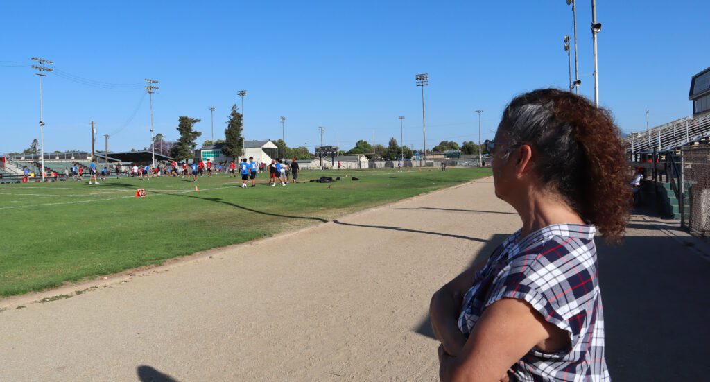 Photo of an older woman with curly white and brown hair wearing a plaid t-shirt and sunglasses stands on a dirt track at an athletic field. Kids are playing flag football on the field in front of her.