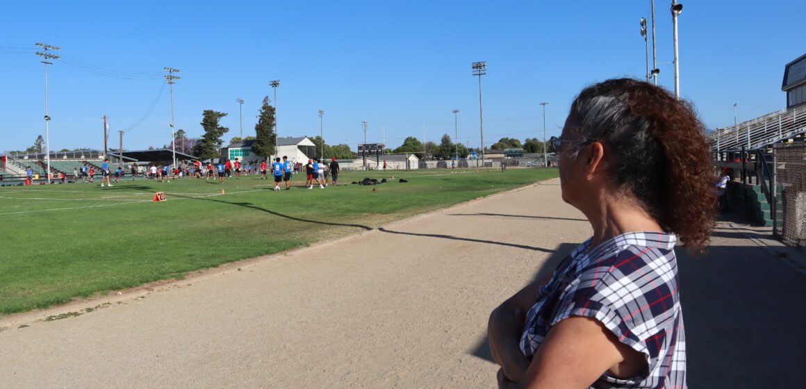 Photo of an older woman with curly white and brown hair wearing a plaid t-shirt and sunglasses stands on a dirt track at an athletic field. Kids are playing flag football on the field in front of her.