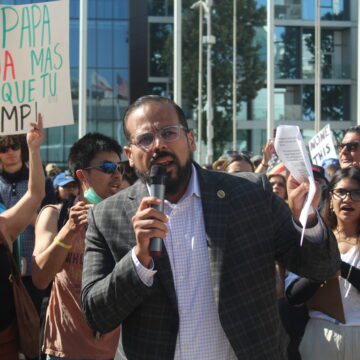 A man loudly proclaiming a speech while holding a mic in one hand and a handful of papers in another.