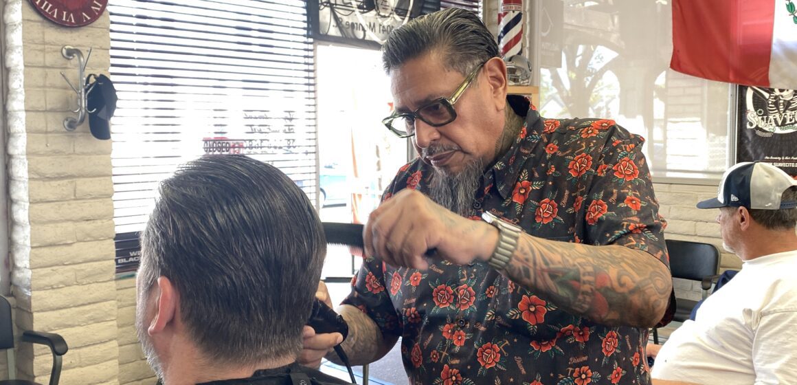Man with slicked back hair and goatee wearing printed shirt combs and trims a customer's hair in a well decorated barbers shop