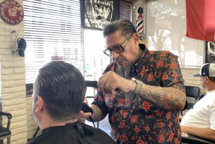 Man with slicked back hair and goatee wearing printed shirt combs and trims a customer's hair in a well decorated barbers shop