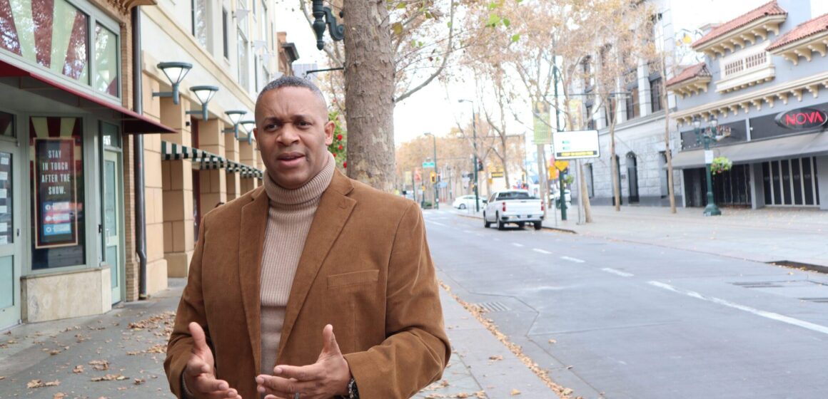 A man in a turtleneck sweater and suit jacket stands outside on the sidewalk next to the street