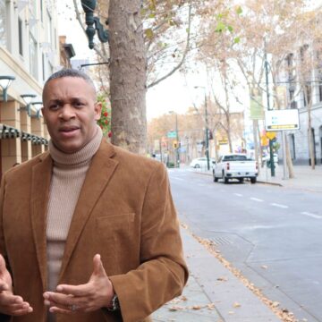 A man in a turtleneck sweater and suit jacket stands outside on the sidewalk next to the street