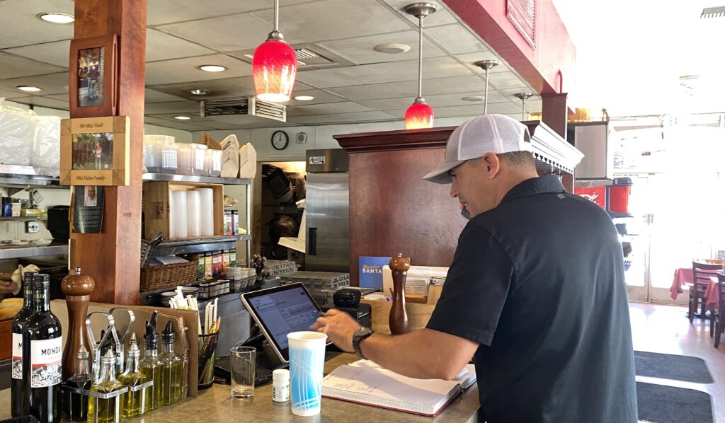 Man with short hair wearing baseball hat talks on phone and plugs in order on screen, with olive oil and other supplies from the Italian restaurant on the counter beside him