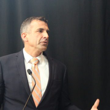 A man in a suit and tie stands at a microphone in front of a black backdrop
