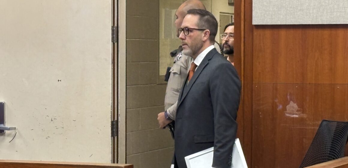 A man in a suit stands in court, with the bailiff and an incarcerated man behind him