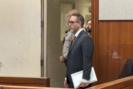 A man in a suit stands in court, with the bailiff and an incarcerated man behind him