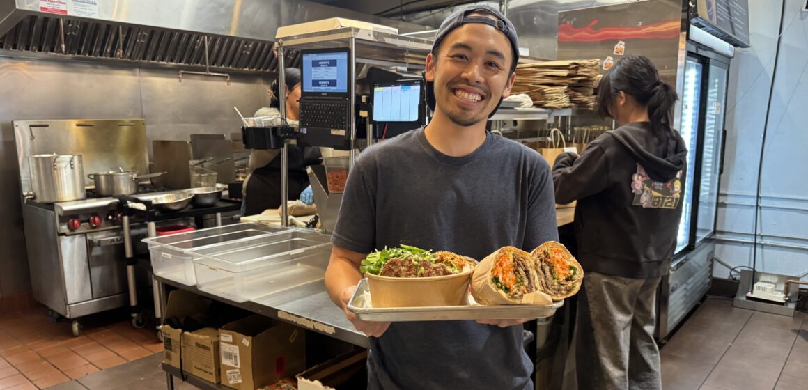 A man in a restaurant kitchen holding a bowl of noodles and a banh mi sandwich