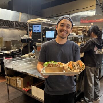 A man in a restaurant kitchen holding a bowl of noodles and a banh mi sandwich