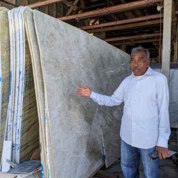 Un hombre mayor con camisa abotonada y jeans señala las losas de granito que hay junto a él.