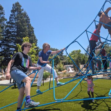 A young man and his mom climb a blue rope play structure