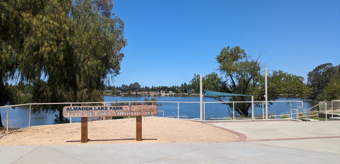 A wooden sign that reads "Almaden Lake Park Terrace Lawn Ampitheater" stands in front of a lake and trees