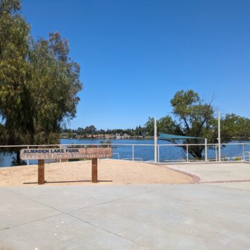 A wooden sign that reads "Almaden Lake Park Terrace Lawn Ampitheater" stands in front of a lake and trees
