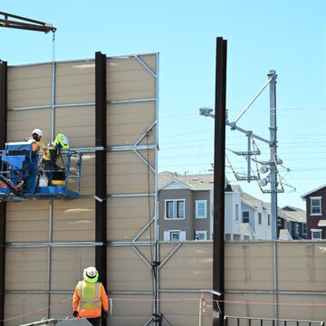 Dos trabajadores en una grúa están trabajando para instalar una pared de paneles, con edificios de apartamentos visibles a través de un espacio en la pared.