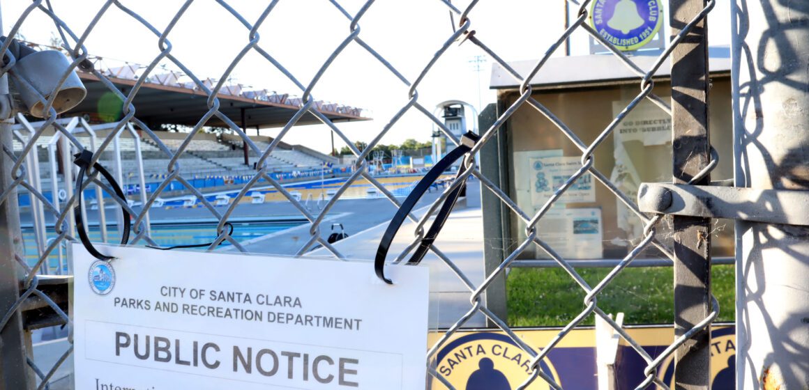 Chainlink fence with posted notice reading "City of Santa Clara Parks and Recreation Department Public Notice International Swim Center Closure." Behind the fence is the Santa Clara Swim Club notice board and two large pools with an audience section.