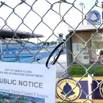 Chainlink fence with posted notice reading "City of Santa Clara Parks and Recreation Department Public Notice International Swim Center Closure." Behind the fence is the Santa Clara Swim Club notice board and two large pools with an audience section.