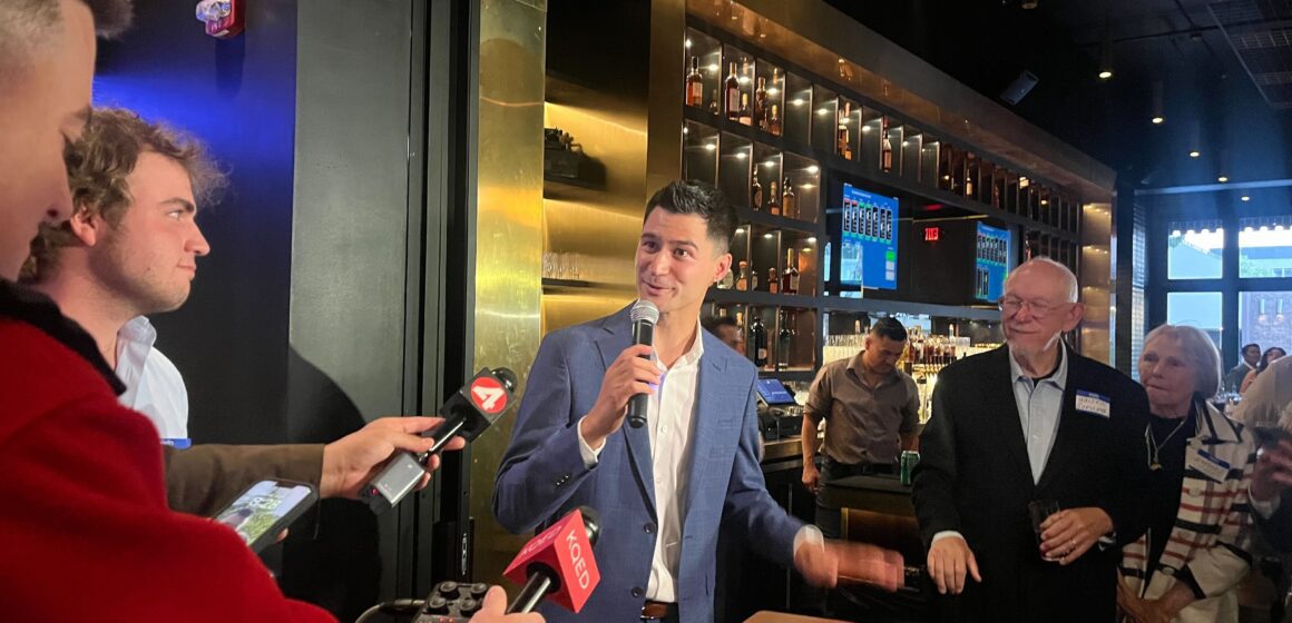 A man in a suit speaking into microphones inside a bar