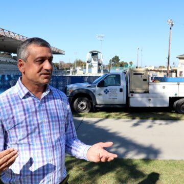 Man wearing plaid shirt talking in front of large pool with construction trucks parked outside