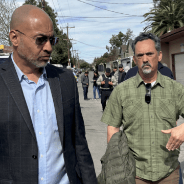 Un hombre con gafas camina junto a otro hombre por una calle larga y concurrida.